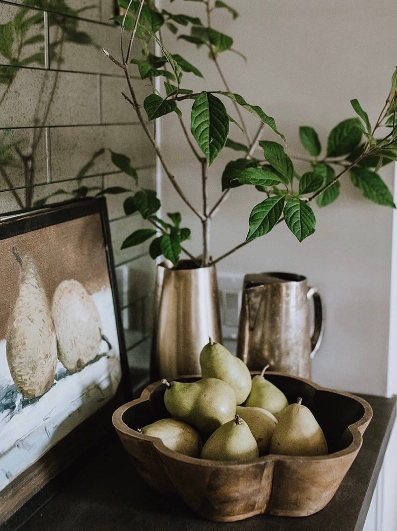 Black Flower Shaped Bowl styled on a countertop, filled with fresh pears, beside a pear painting and brass pitchers