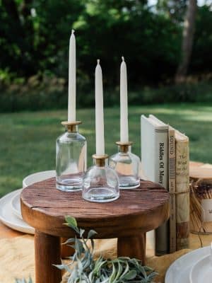 A trio of Skinny Taper Glass Candle Holders with white taper candles, displayed on a Round Wood Pedestal alongside a Decorative Vintage Books Set – Neutral Colors in a natural outdoor setting.