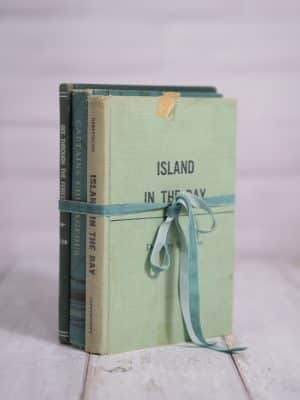 Three hardback books in cool green and blue tones, tied with teal ribbon, upright on a rustic white background