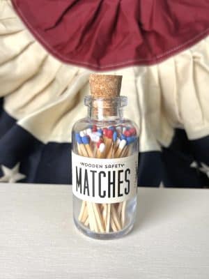 Small glass jar filled with red, white, and blue tip matches, sealed with a cork top and labeled “Wooden Safety Matches,” placed in front of Americana bunting.