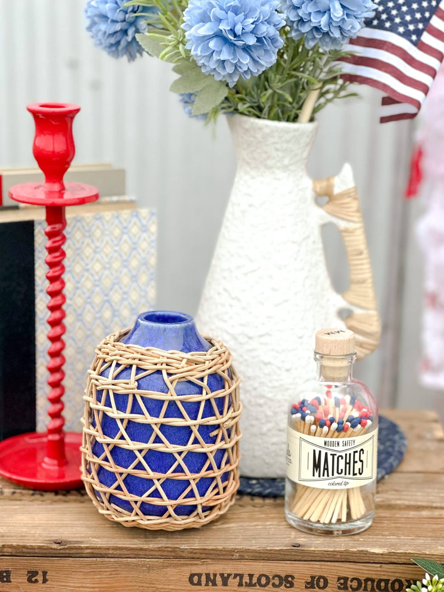 Patriotic vignette with red candle holder, indigo rattan vase, large white rattan-handled vase, and vintage-style matches.