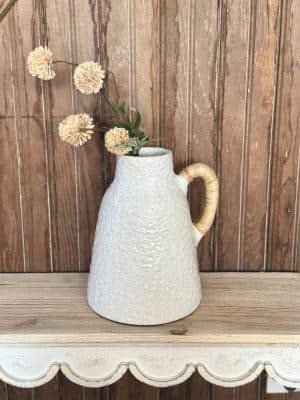 Small ceramic vase with speckled off-white glaze and woven rattan handle, styled with dried flowers on a scalloped shelf.