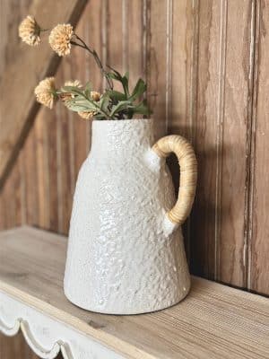 Ceramic vase with textured off-white glaze and rattan-wrapped handle, styled with dried flowers on a rustic wood shelf.
