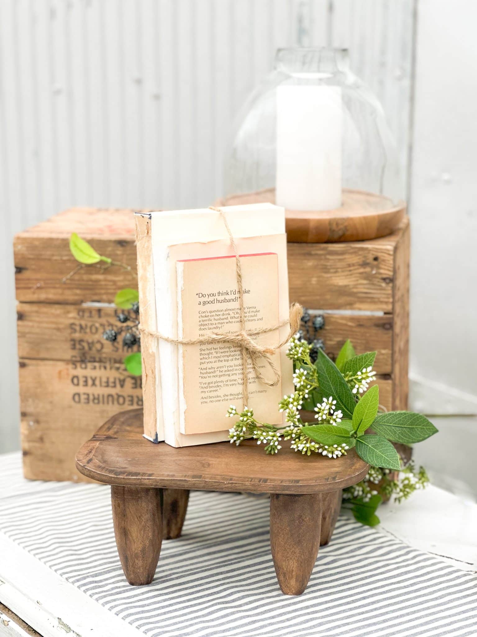 Decorative vintage softcover books tied with twine, styled on a small carved wood stool with white florals, beside a large wood and glass hurricane candle holder on a rustic wood crate.