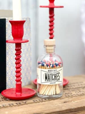 Close-up of Vintage Apothecary USA Matches in a clear glass jar with cork top, flanked by red candle holders and vintage books.