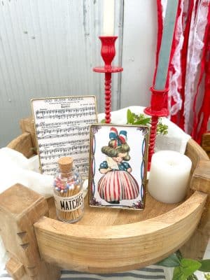 Close-up of a patriotic vignette on a wood chakki table pedestal, featuring two red corkscrew taper holders, framed God Bless America sheet music and a vintage girl illustration in gold industrial frames, a white candle, and a jar of red, white, and blue wooden matches.