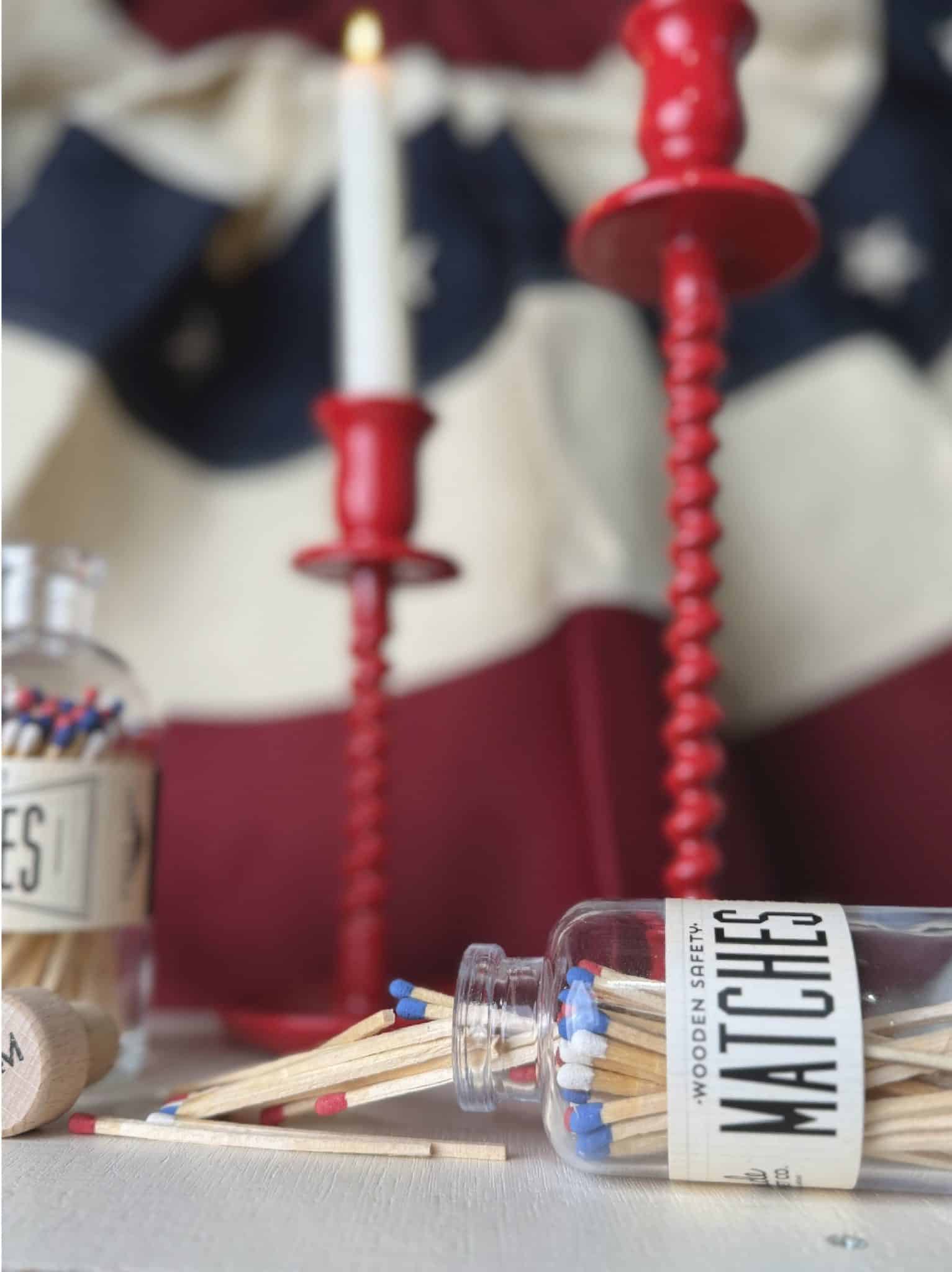 Patriotic scene with spilled mini USA match jar and full match jar in the background, styled alongside red corkscrew candle holders and a lit white taper.