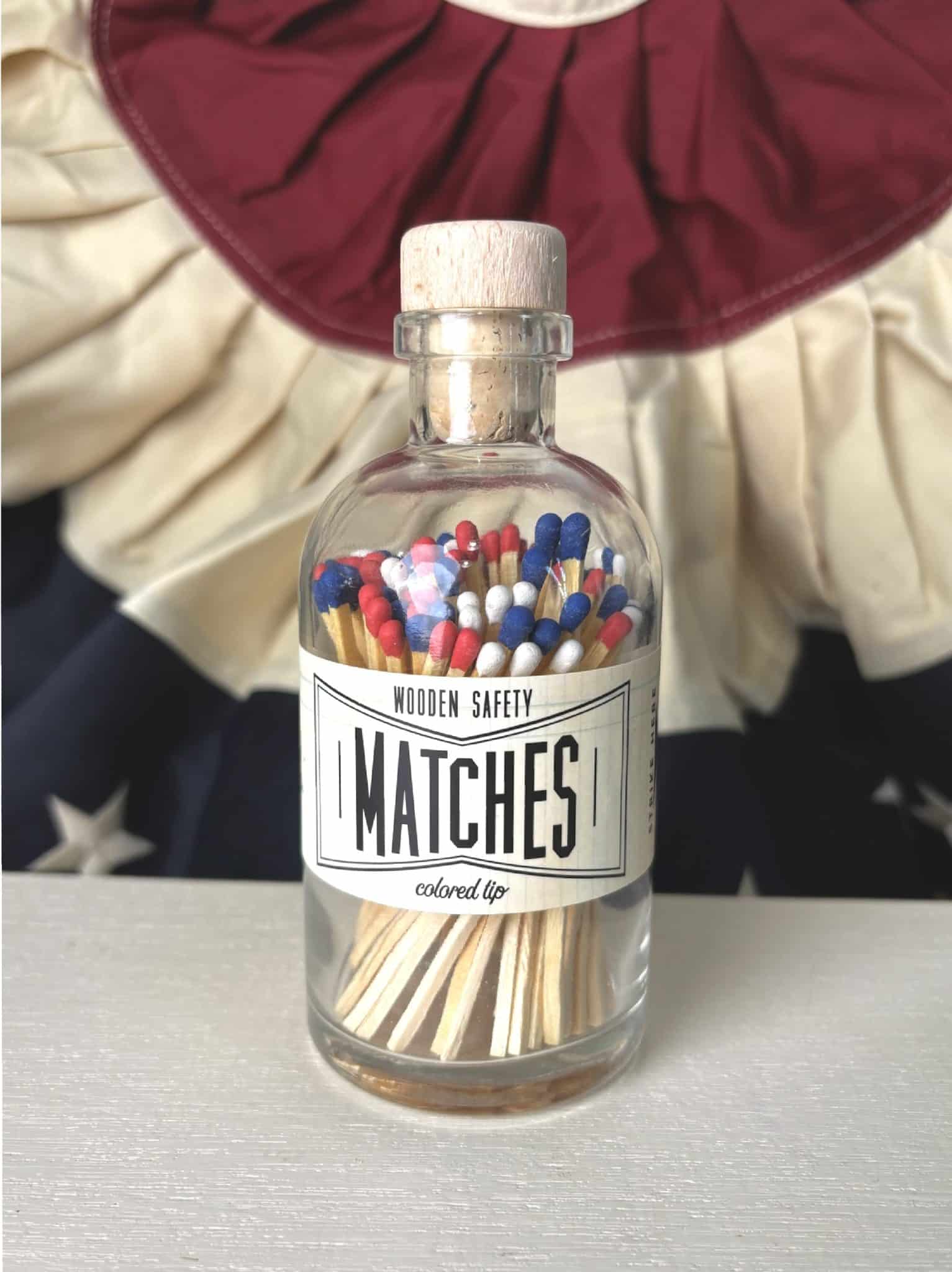 Apothecary-style match jar filled with tri-color matchsticks and labeled “Wooden Safety Matches,” placed in front of a flag-inspired backdrop.