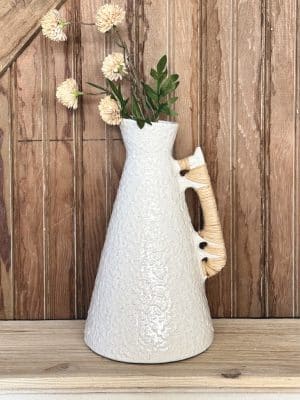 Ceramic vase with speckled white glaze and woven rattan handle, styled with faux florals on a scalloped-edge shelf.