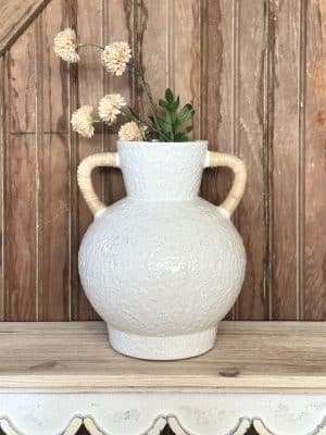 Ceramic vase with a rounded silhouette and two rattan-wrapped handles, styled with faux florals against wood paneling.