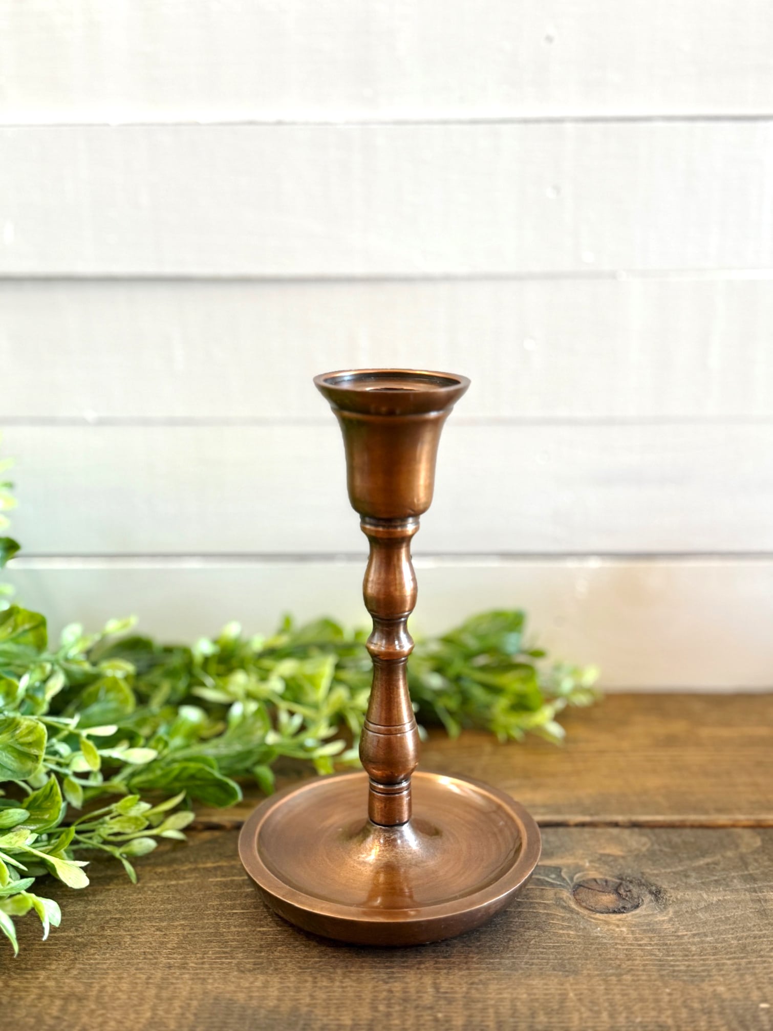 Antique copper taper candle holder in small size displayed on a wood surface with greenery in background.