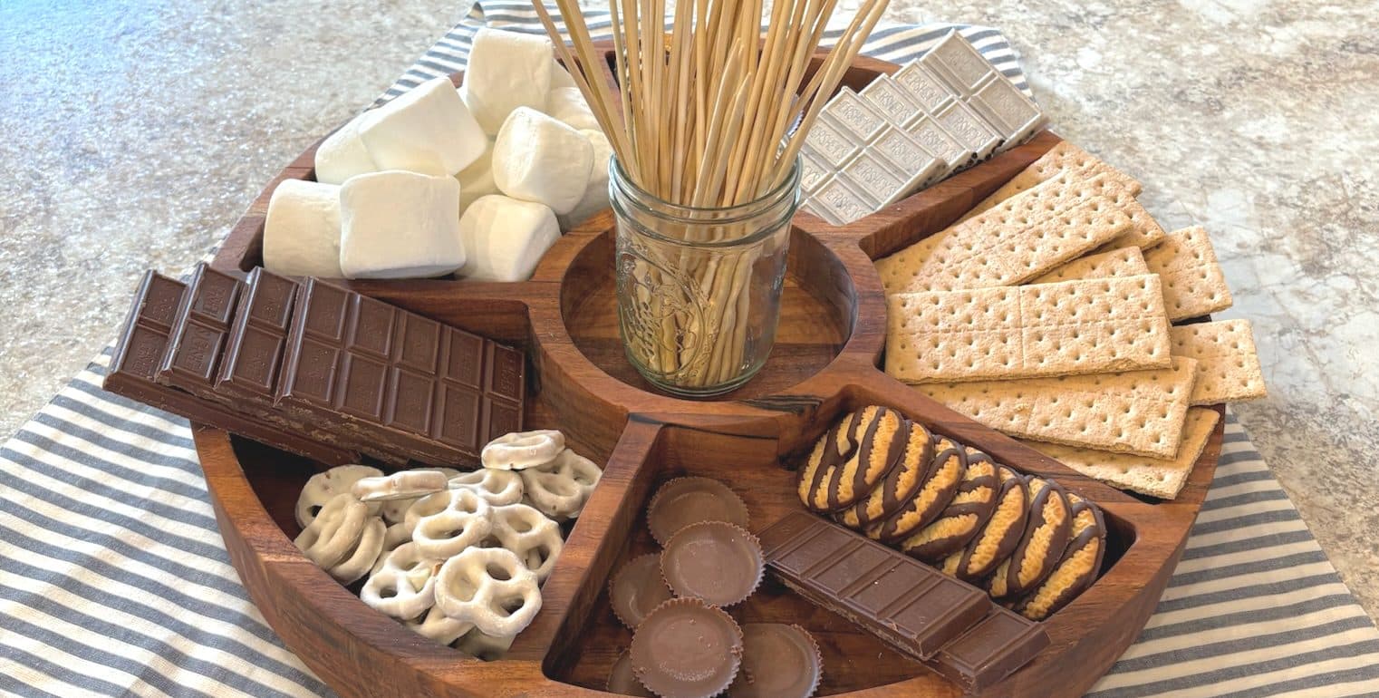 Divided wooden tray filled with s’mores ingredients—chocolates, graham crackers, marshmallows, and pretzels—styled on a striped cloth.