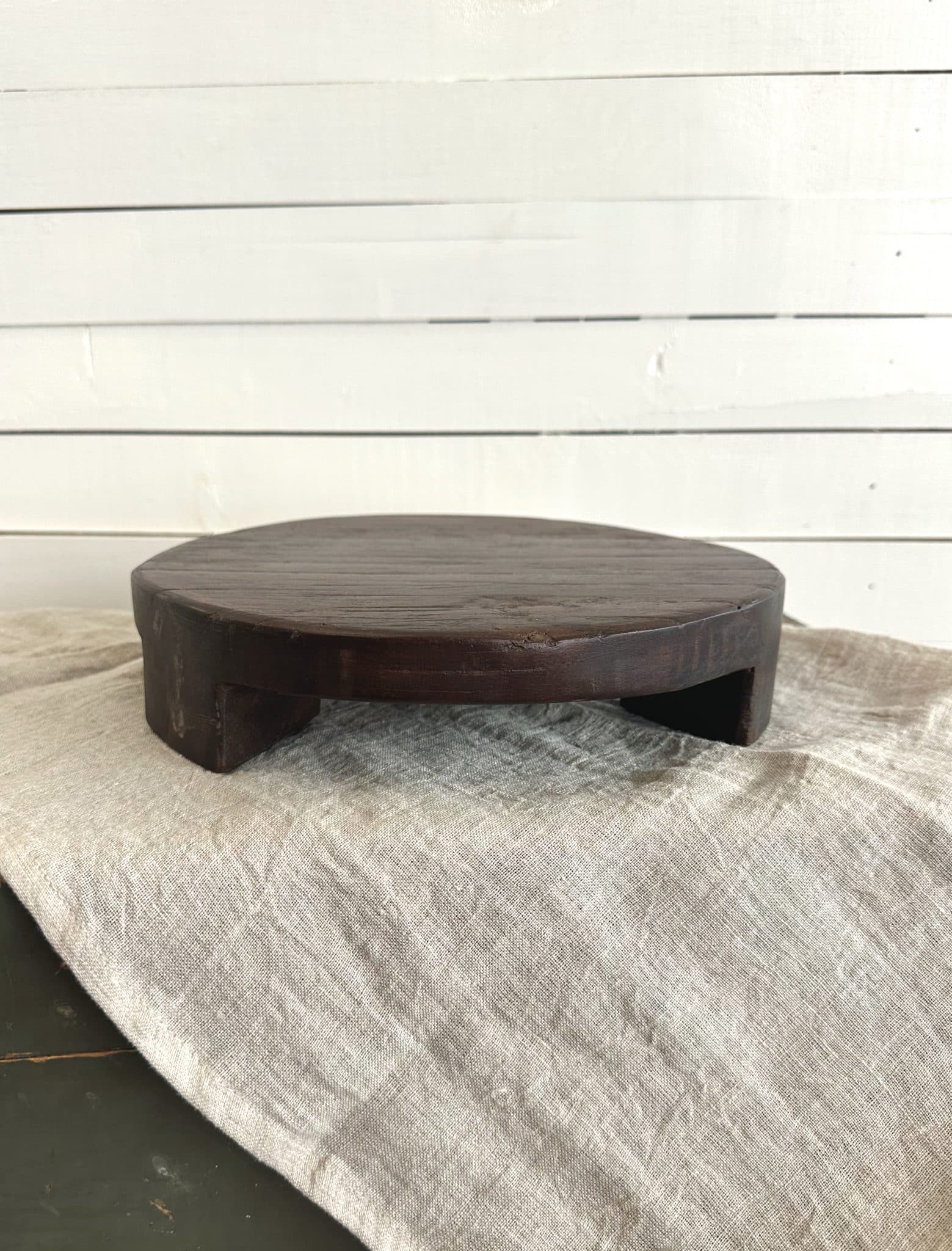 Medium-sized round wood pedestal with short feet and dark stain, displayed against a white shiplap backdrop.