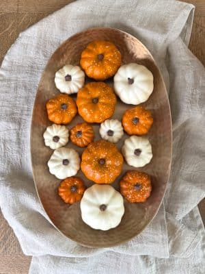 Top-down view of an oval brass platter filled with alternating mini white and orange pumpkins, styled on a soft neutral cloth.