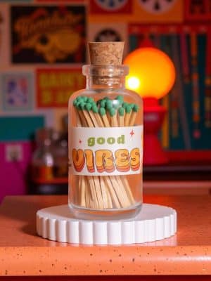 Glass apothecary jar filled with green-tipped matches labeled “Good Vibes,” styled on a retro orange and pink background with warm lighting.