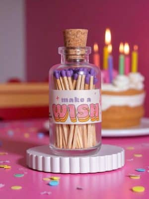 Glass apothecary jar of purple-tipped matches labeled “Make a Wish,” styled with a pink background and birthday cake with candles.
