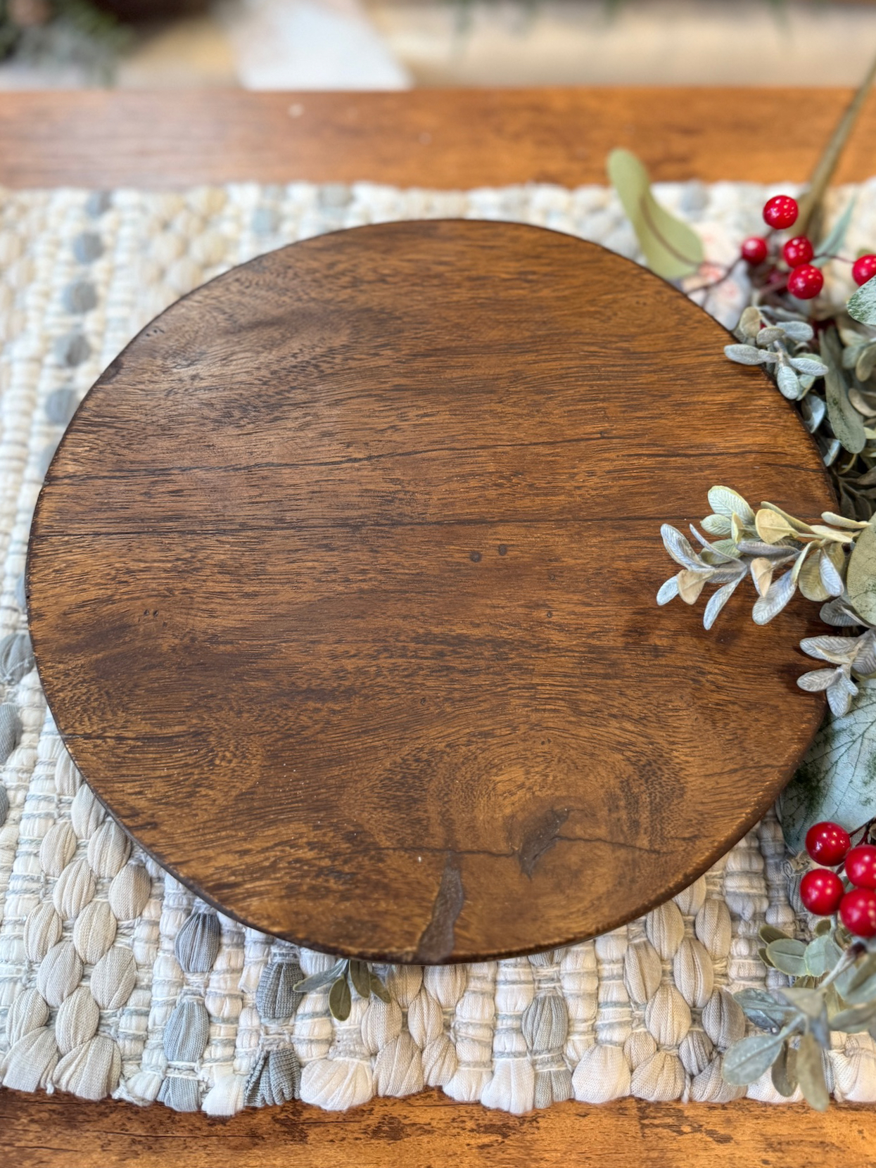 Found wood pedestal with warm brown grain, styled on a woven mat with winter greenery and red berries.