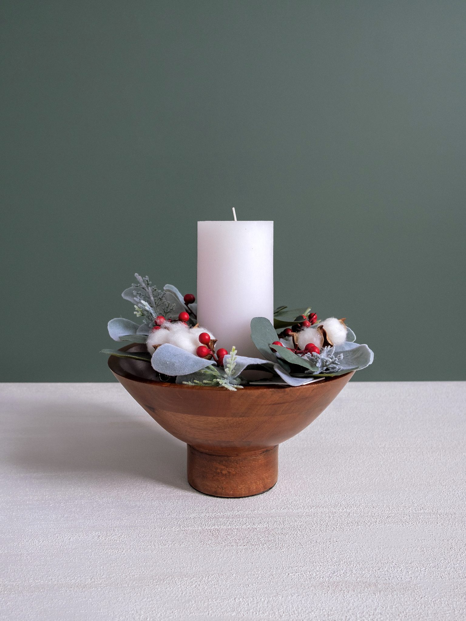 Mini cotton and eucalyptus Christmas candle wreath styled around a white pillar candle inside a wooden pedestal bowl.