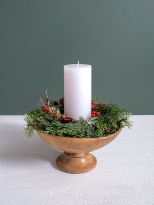 Classic wood pedestal bowl styled with winter greenery and a white pillar candle on a white tabletop.