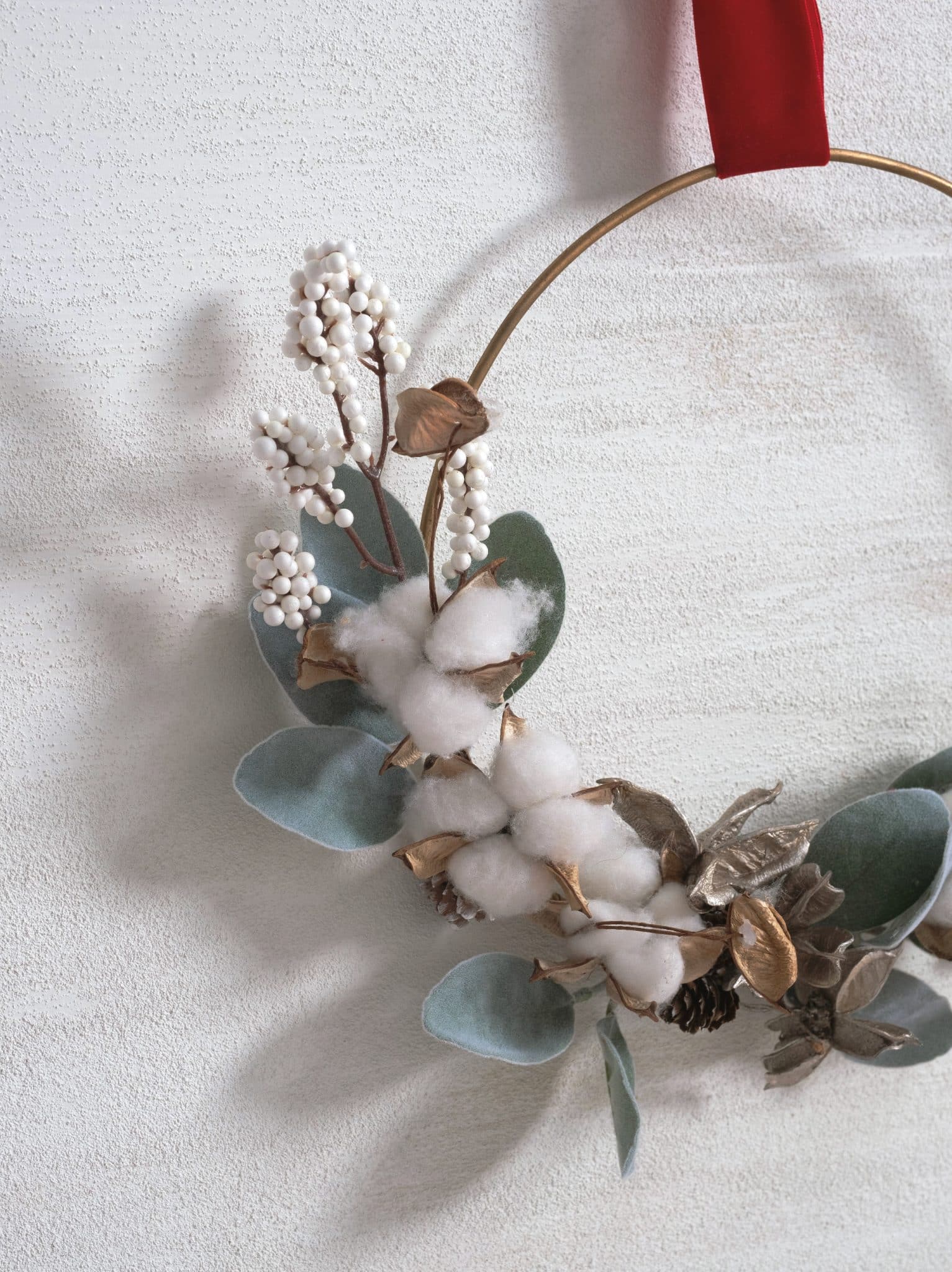 Gold metal Christmas wreath ring with faux eucalyptus, cotton stems, white berries, and a red velvet ribbon hanging on top.