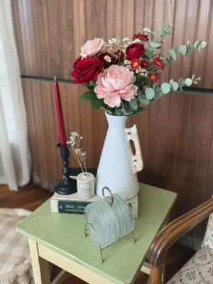 Large off-white ceramic vase with rattan-wrapped handle, filled with red and blush flowers, styled on a green side table with a black candle holder and stacked books.