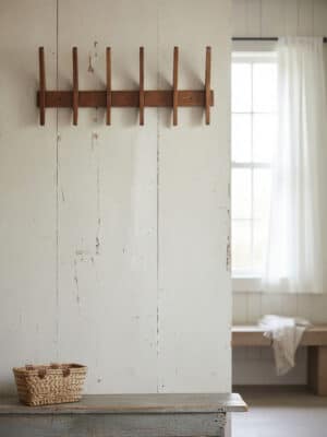 Large wood hanging wall rack with five curved pegs mounted on a white plank wall near a window.