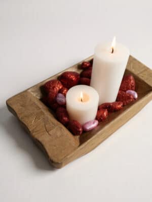 Small hand-carved wooden dough bowl with white pillar candles, red berries, and candy hearts styled as a cozy centerpiece.