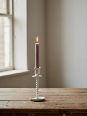 Off-white cast iron corkscrew taper candle holder small with a lit taper candle styled on a rustic wooden table near a window.